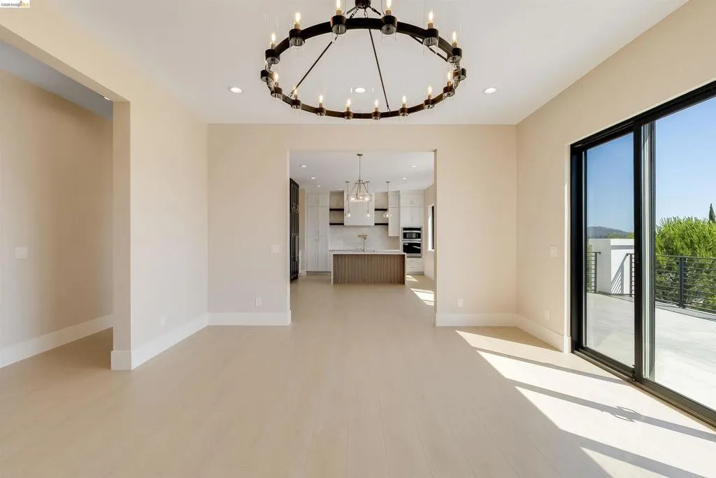 Open-concept living area featuring light wood-finish flooring, a large circular chandelier, recessed lighting, and a sliding glass door opening to an outdoor patio with a metal railing