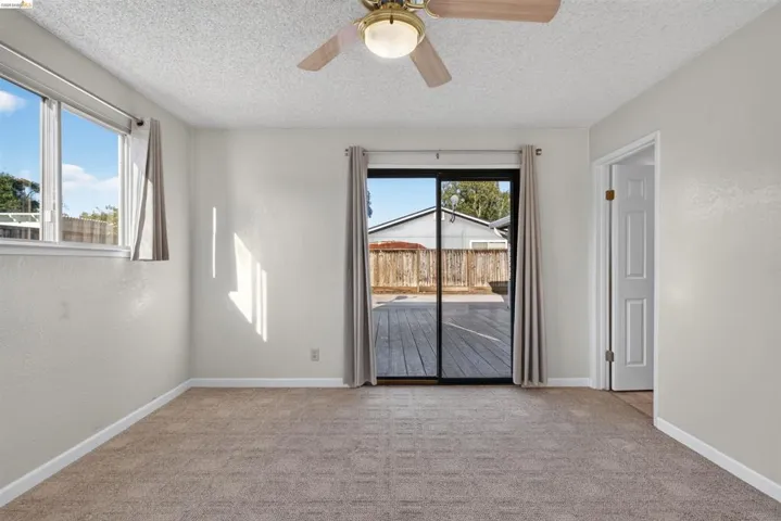 Neutral-toned room with plush carpeting, featuring a ceiling fan with integrated lighting, and a sliding glass door opening to an outdoor deck