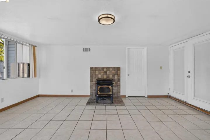 Spacious room featuring neutral-toned tile flooring, a wood-burning stove with a tiled surround, and multiple window and door openings