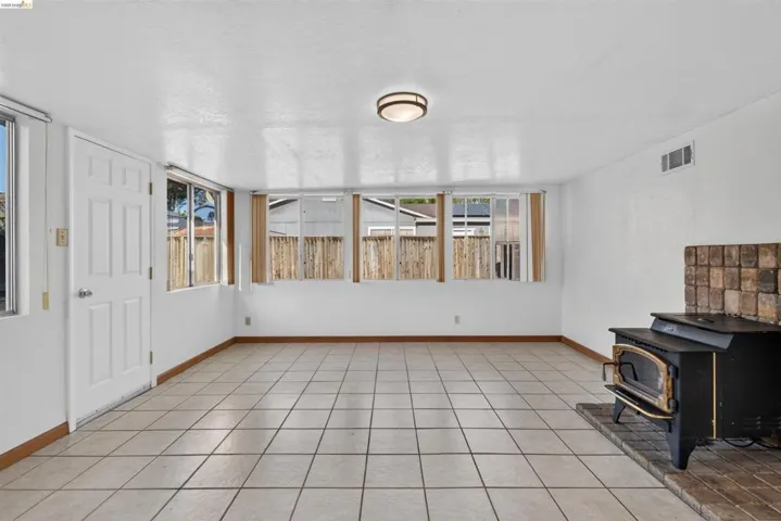 Spacious room featuring tile flooring, multiple windows, a white paneled door, wood-burning stove with a brick hearth, and a flush-mount ceiling light fixture