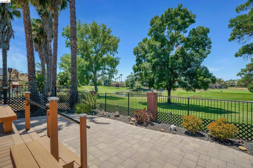 Paver patio with a black metal fence, mature palm trees, and golf course landscaping featuring a pond and trees