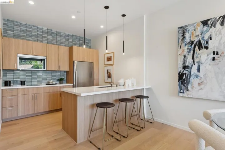 Modern kitchen featuring natural wood cabinetry, a light-toned wood-finish floor, a stainless steel refrigerator, and a tiled backsplash with blue and grey tones