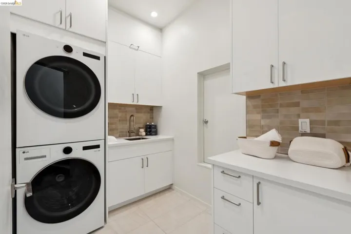 Laundry room featuring stacked front-load machines, extensive cabinetry with brushed nickel hardware, and a utility sink