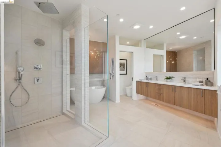 Spacious bathroom featuring a glass-enclosed shower with a rain shower head and handheld sprayer, a freestanding soaking tub, a floating wood-finish double vanity with dual sinks, and a large wall-mounted mirror