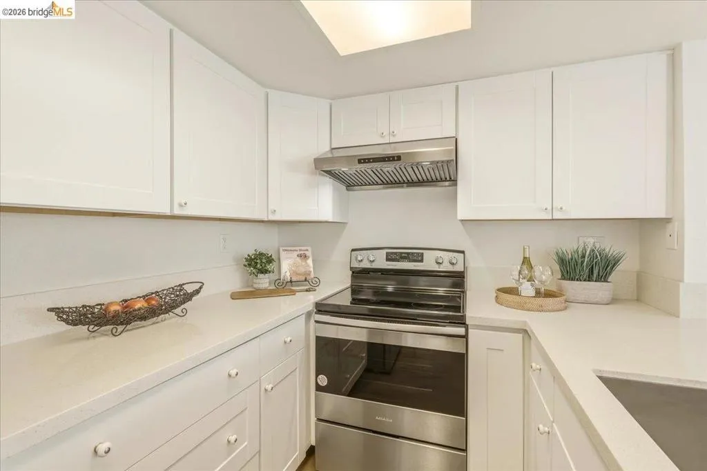 U-shaped kitchen featuring white shaker cabinetry, light-toned countertops, a stainless steel range with matching hood, and an undermount sink U-shaped kitchen featuring white shaker cabinetry, light-toned countertops, a stainless steel range with matching hood, and an undermount sink