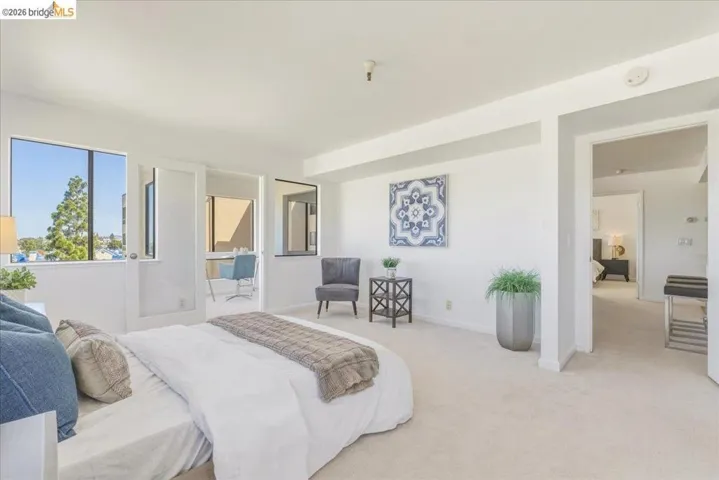 Open-concept bedroom featuring large windows, light-colored carpeting, an adjoining flexible space, and a doorway opening to an additional room