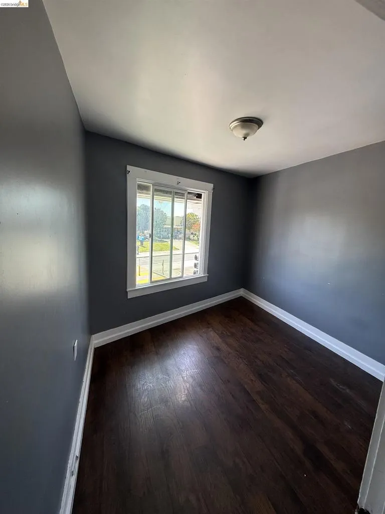 Compact room featuring rich wood-finish flooring, crisp white baseboards, and a single window with white trim