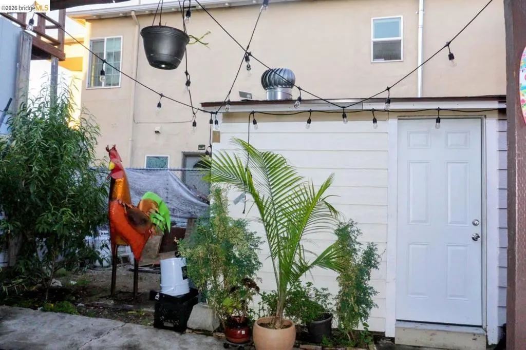 White paneled exterior door with a window unit above, featuring string lighting, potted plants, and concrete paving