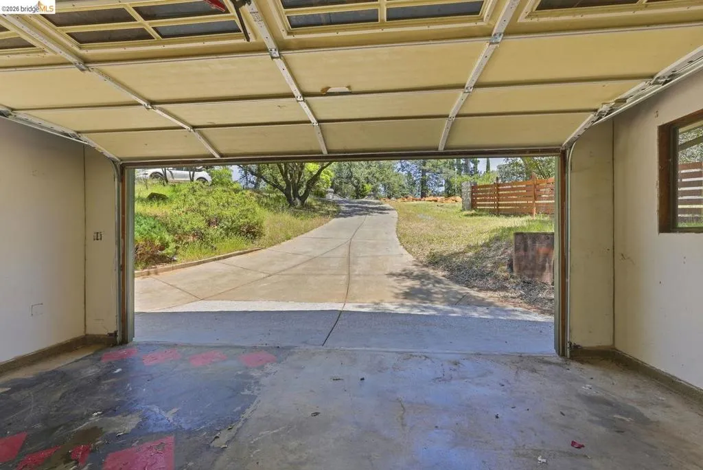 Open garage with concrete flooring, showcasing a concrete driveway leading to a treed landscape