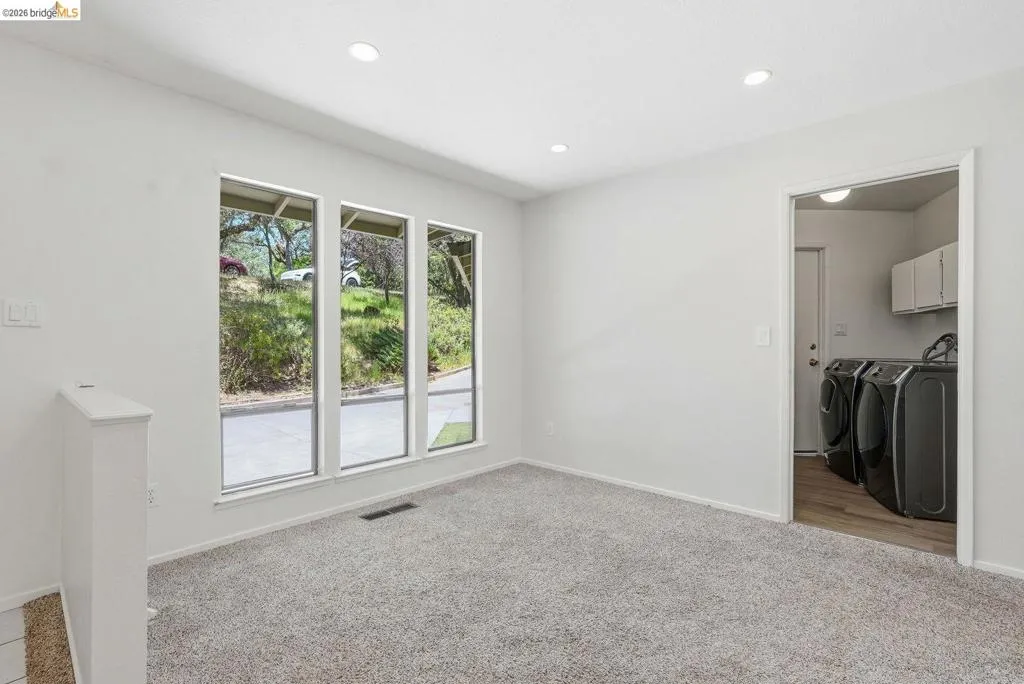 Bright room featuring three large windows, recessed ceiling lighting, light-colored carpeting, and a dedicated laundry room entrance with wood-finish flooring