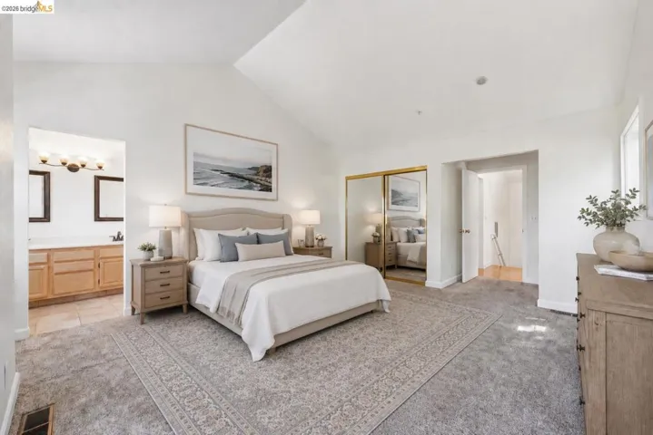 Vaulted ceiling bedroom with plush carpeting, featuring a mirrored closet, and an en-suite bathroom with a double vanity and tile flooring