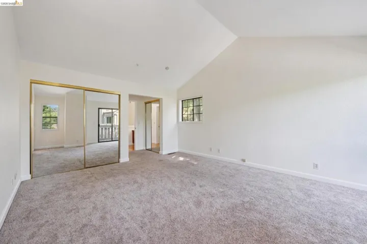 Spacious room featuring vaulted ceilings, neutral-toned carpeting, mirrored closet doors with gold-tone frames, and multiple windows providing natural illumination