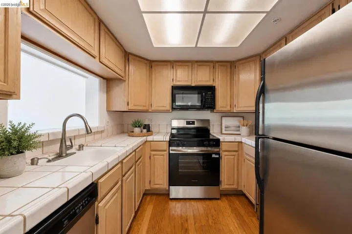 U-shaped kitchen featuring light wood cabinetry, tile countertops and backsplash, stainless steel appliances, and wood-finish flooring