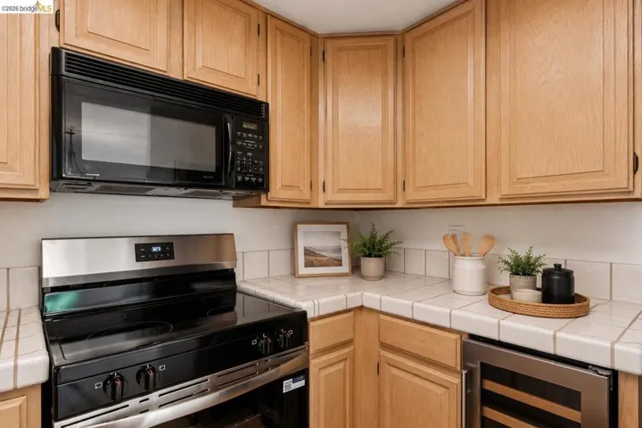 Wood-finish cabinetry and tiled countertops with an integrated stainless steel wine cooler, black electric range, and coordinating microwave