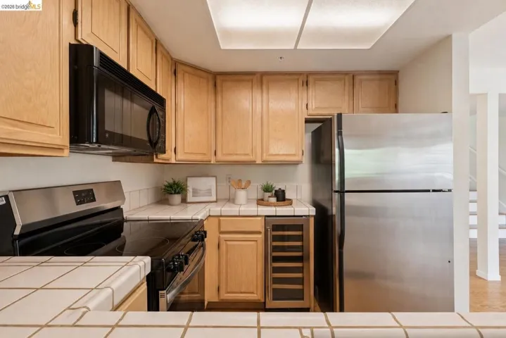 Kitchen featuring light wood cabinetry, white tile countertops, stainless steel refrigerator, black range, and built-in microwave