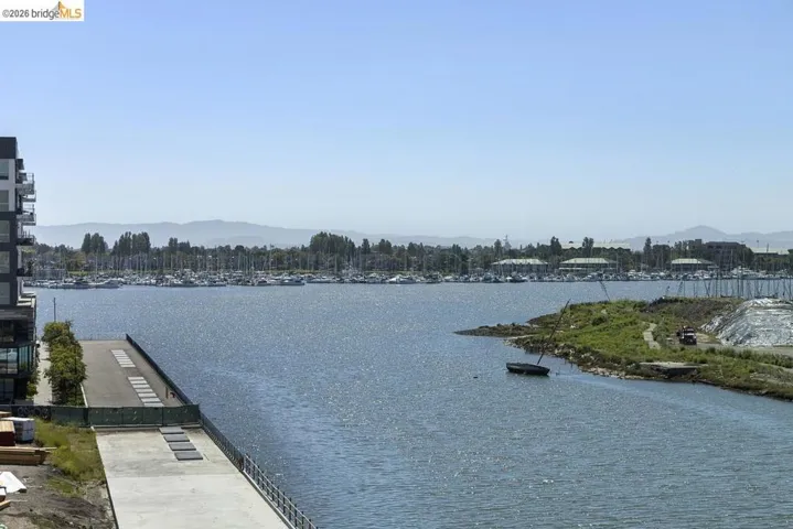 Waterfront setting featuring a modern building with balconies, a marina with numerous boats, a paved promenade, and distant mountains