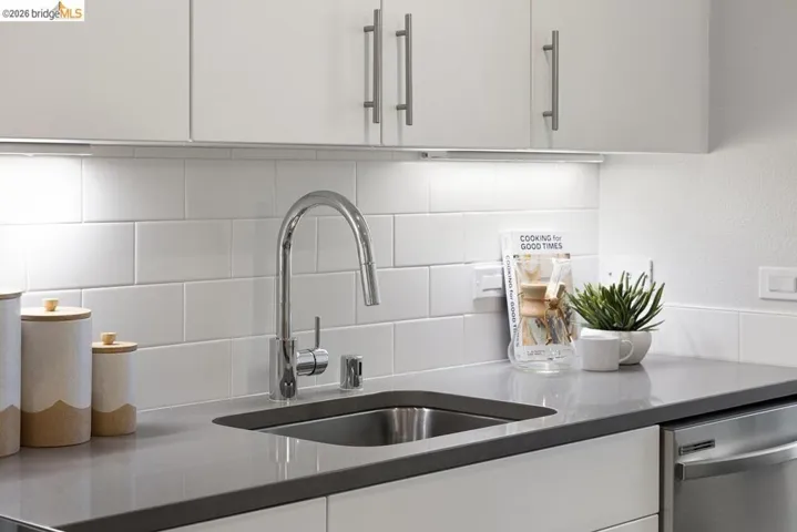 Kitchen featuring a stainless steel sink, chrome gooseneck faucet, gray countertop, white subway tile backsplash, white upper and lower cabinetry with brushed metal hardware, and under-cabinet lighting