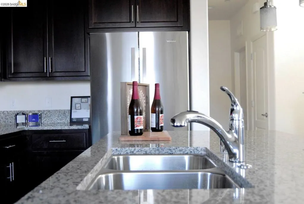 Kitchen featuring a double-basin stainless steel sink with a chrome gooseneck faucet, granite countertops, dark wood cabinetry with brushed nickel hardware, and a stainless steel refrigerator