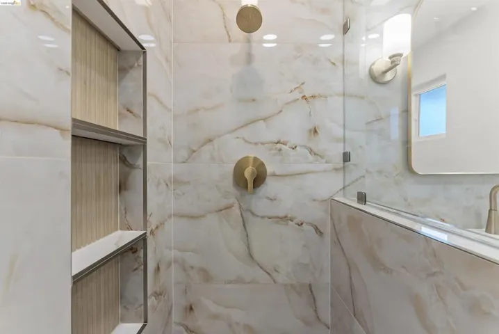 Shower enclosure featuring floor-to-ceiling marbled-patterned large format tile, brushed gold shower fixtures, a clear glass shower door, and a built-in shower niche with ribbed wood-finish backing