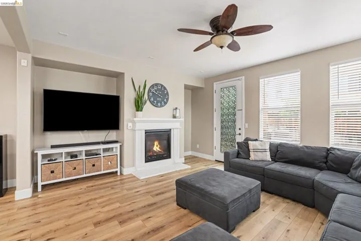 Spacious living area featuring wood-finish flooring, a white mantel fireplace, ceiling fan, and multiple windows with blinds