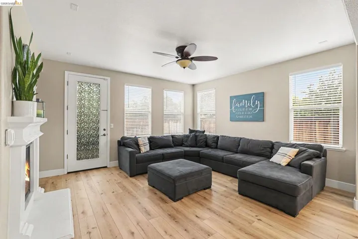 Spacious living area featuring wood-finish flooring, multiple windows with blinds, a white fireplace with mantle, a ceiling fan with integrated lighting, and a door with frosted glass