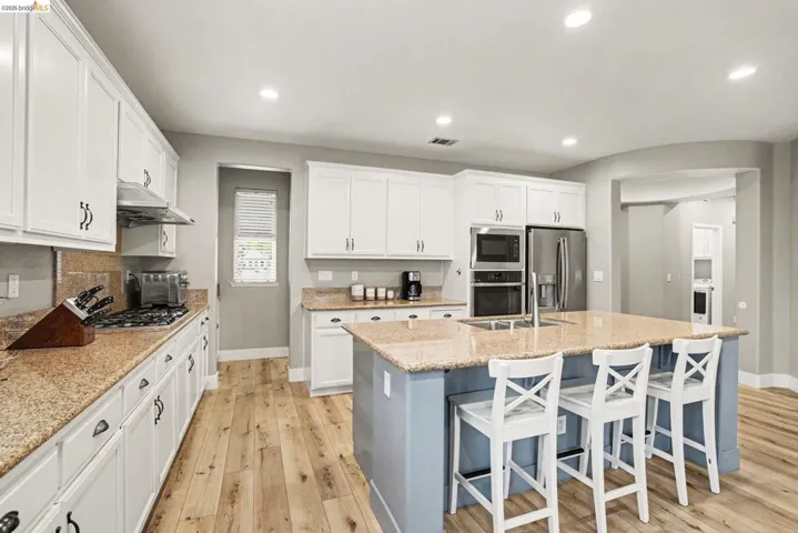 Spacious kitchen featuring an expansive island with seating, abundant white cabinetry, granite countertops, wood-finish flooring, and stainless steel appliances