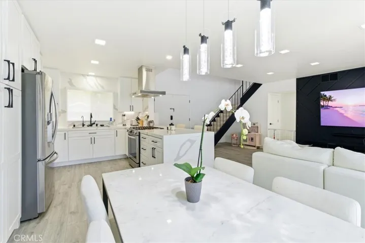 Open-concept kitchen and dining area featuring white cabinetry, stainless steel appliances, a marble dining table, and modern lighting, flowing seamlessly into the living room with views of the staircase and black accent wall.