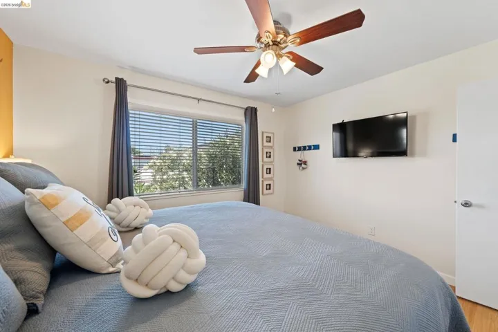 Bedroom featuring a ceiling fan with light fixture, window with blinds and dark drapery, wall-mounted television, and wood-finish flooring