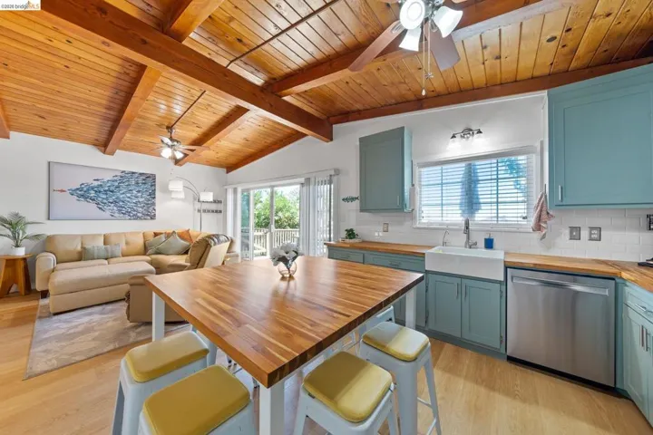 Open-concept living space featuring a vaulted wood-paneled ceiling with exposed beams, wood-finish flooring, a kitchen with teal cabinetry, butcher block countertops, and a farmhouse sink, and sliding glass doors opening to an exterior deck