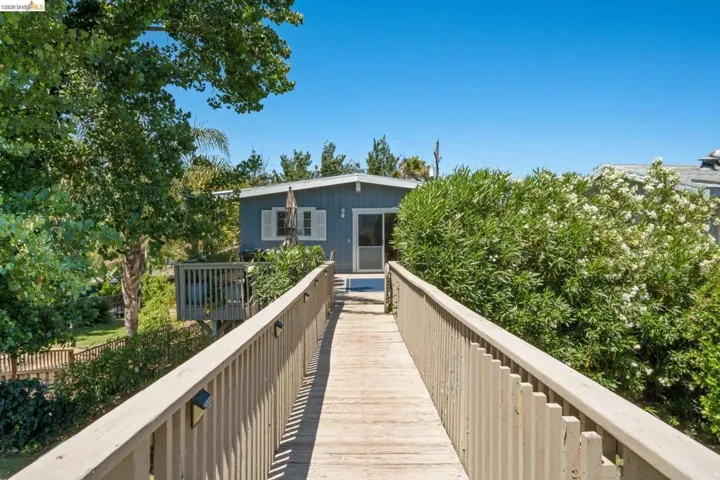 Elevated wood-finish walkway providing access to a property featuring a light blue exterior, white window shutters, and a sliding glass door