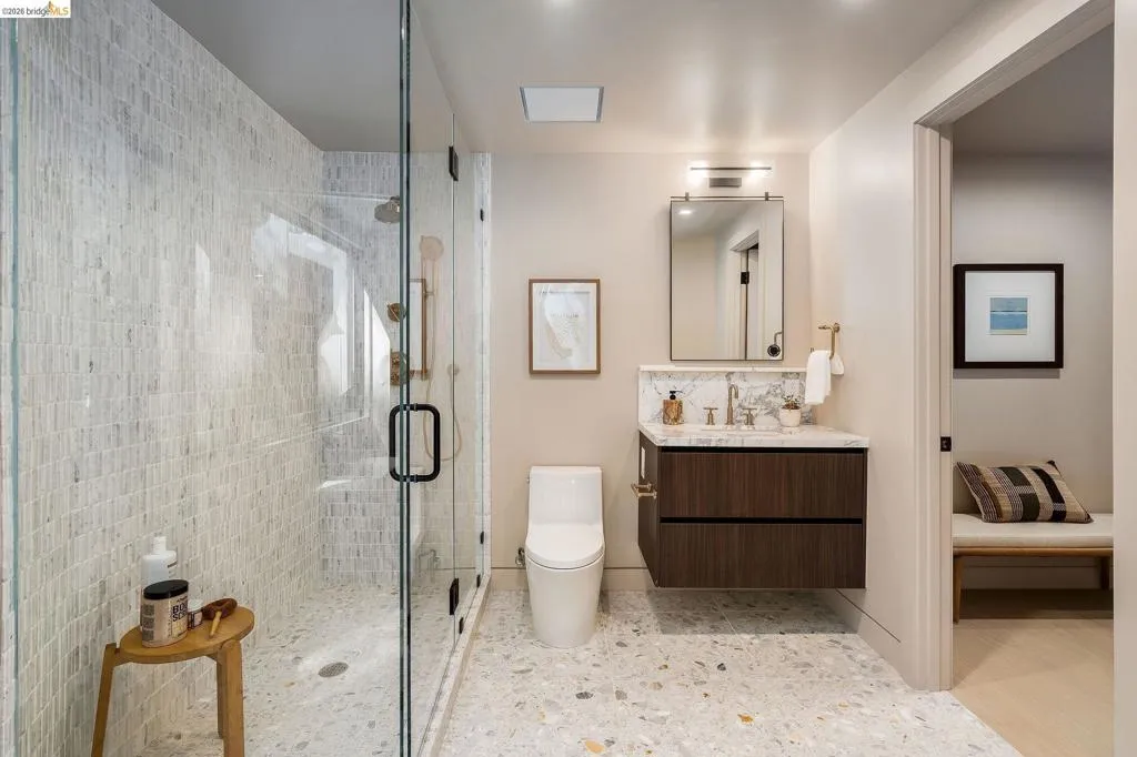 Spacious bathroom featuring a glass-enclosed shower with mosaic wall tile, a floating wood-finish vanity with a stone countertop, a rectangular mirror with integrated lighting, and terrazzo-style flooring