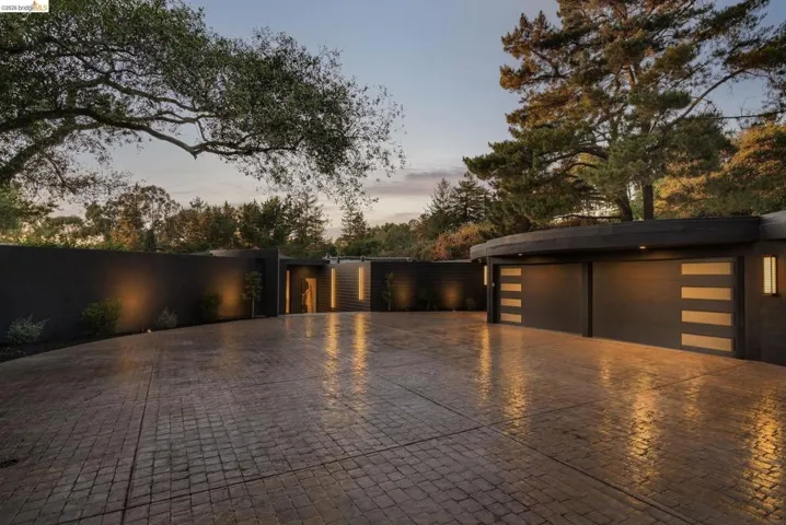 Expansive stamped concrete driveway with custom exterior lighting, a curved architectural garage featuring two doors with frosted panel inserts, and a prominent solid privacy wall