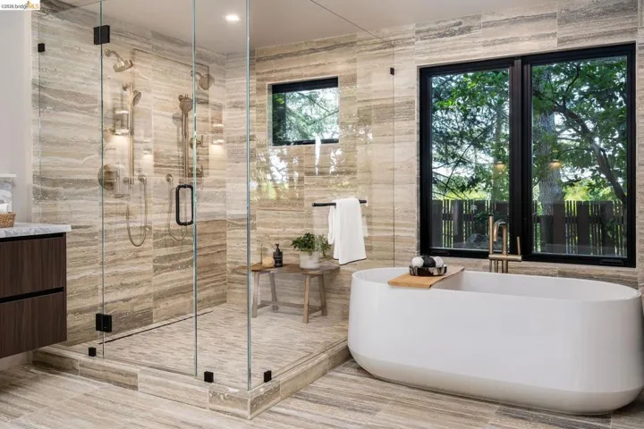 Spacious bathroom featuring a frameless glass shower enclosure with multiple showerheads, a freestanding soaking tub with floor-mounted fixtures, and a dual-pane window with black trim