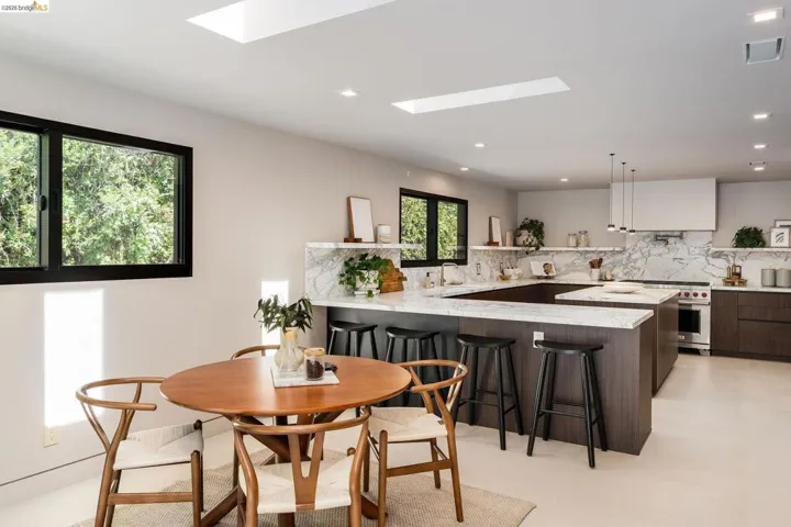 Modern kitchen featuring a large central island with seating, dark wood-finish cabinetry, stone countertops with matching backsplash, stainless steel appliances, and skylights providing abundant overhead illumination