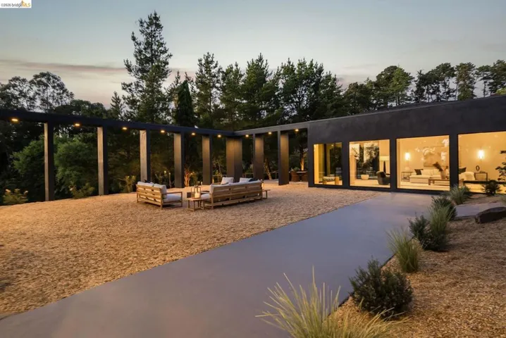 Modern single-story exterior featuring a dark facade with expansive windows, a gravel patio, and a covered pergola with integrated lighting