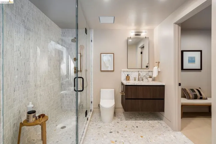 Spacious bathroom featuring a glass-enclosed shower with mosaic wall tile, a floating wood-finish vanity with a stone countertop, a rectangular mirror with integrated lighting, and terrazzo-style flooring