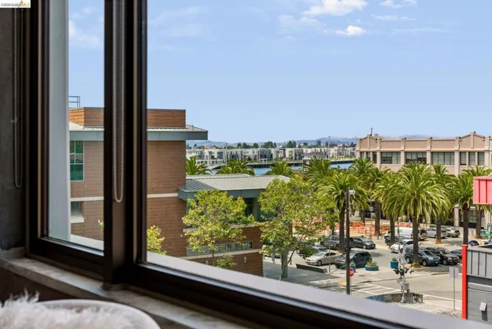 Expansive framed window overlooking a vibrant urban streetscape with palm trees, waterfront buildings, and distant hills at the entrance to Jack London Square