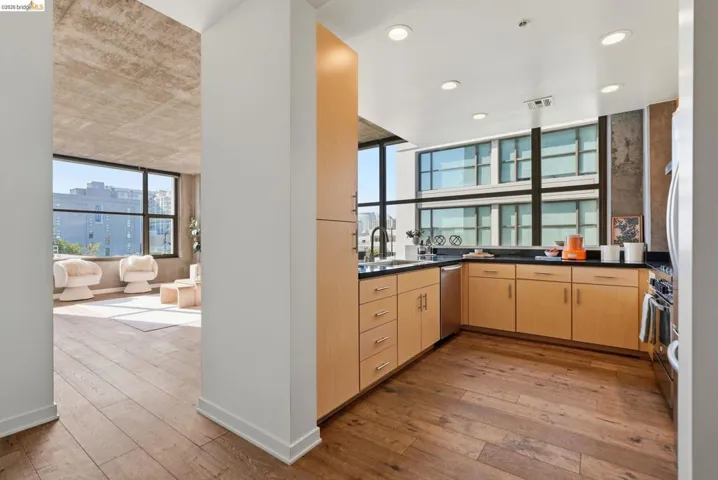 Open-concept kitchen with wood-finish flooring, light wood cabinetry, black countertops, and stainless steel appliances