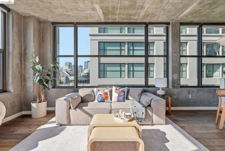 Expansive living area featuring floor-to-ceiling windows, exposed concrete walls and ceiling, wood-finish flooring, and a light-toned area rug