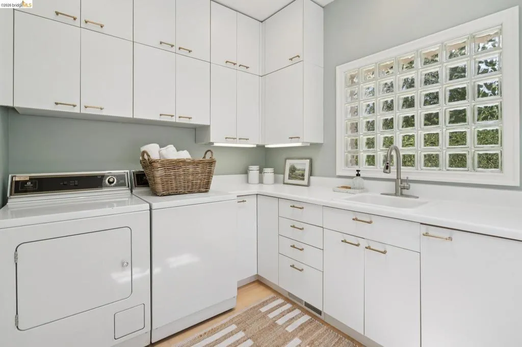 Laundry room featuring white cabinetry with gold-tone hardware, a single basin sink with a chrome faucet, light wood-finish flooring, and a glass block window