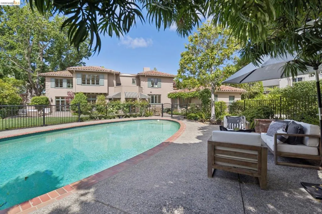 Curved swimming pool with brick coping, surrounded by a concrete patio and a black wrought iron fence