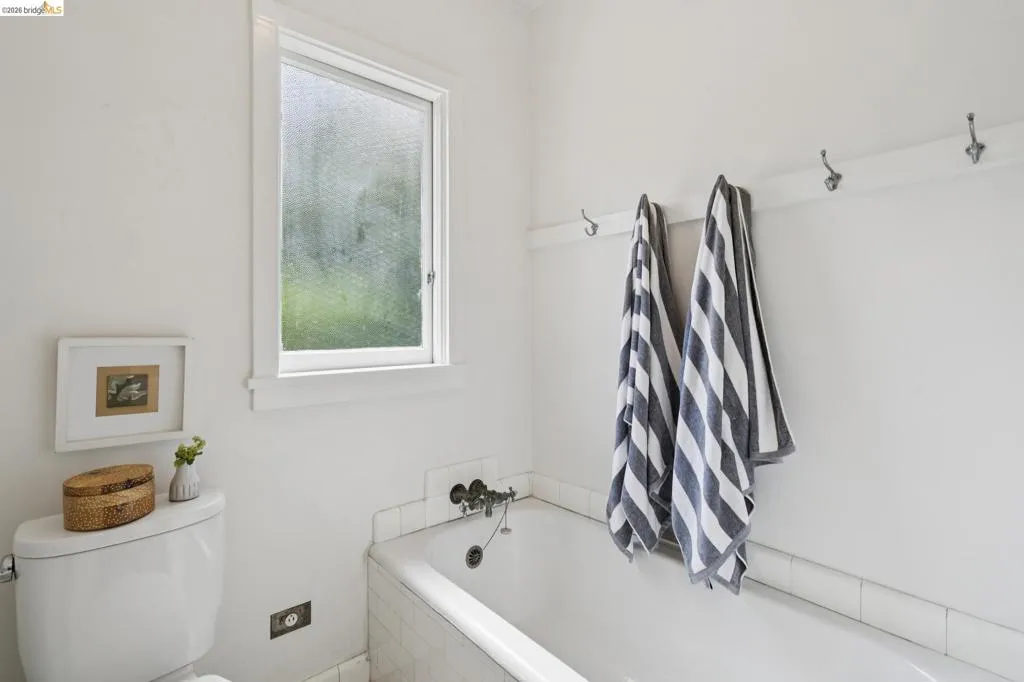 Bathroom featuring a white bathtub with tile surround, a privacy window, wall-mounted towel hooks, and a white toilet