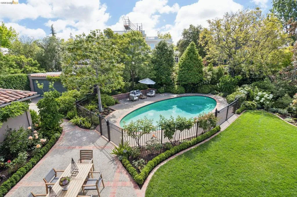 Expansive exterior featuring a freeform swimming pool, surrounding wrought iron fencing, a spacious paver patio, a separate lawn area, and mature landscaping with established trees and shrubs