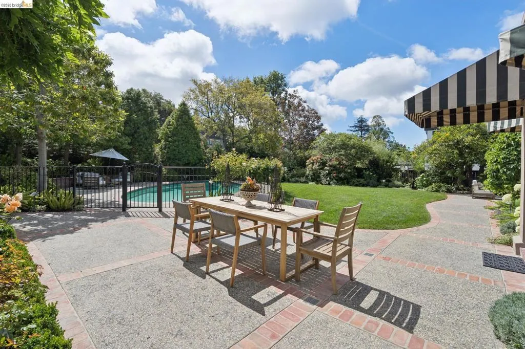 Expansive backyard featuring a manicured lawn, a swimming pool with a black wrought iron fence, a stone patio with brick accents, and mature trees and shrubs