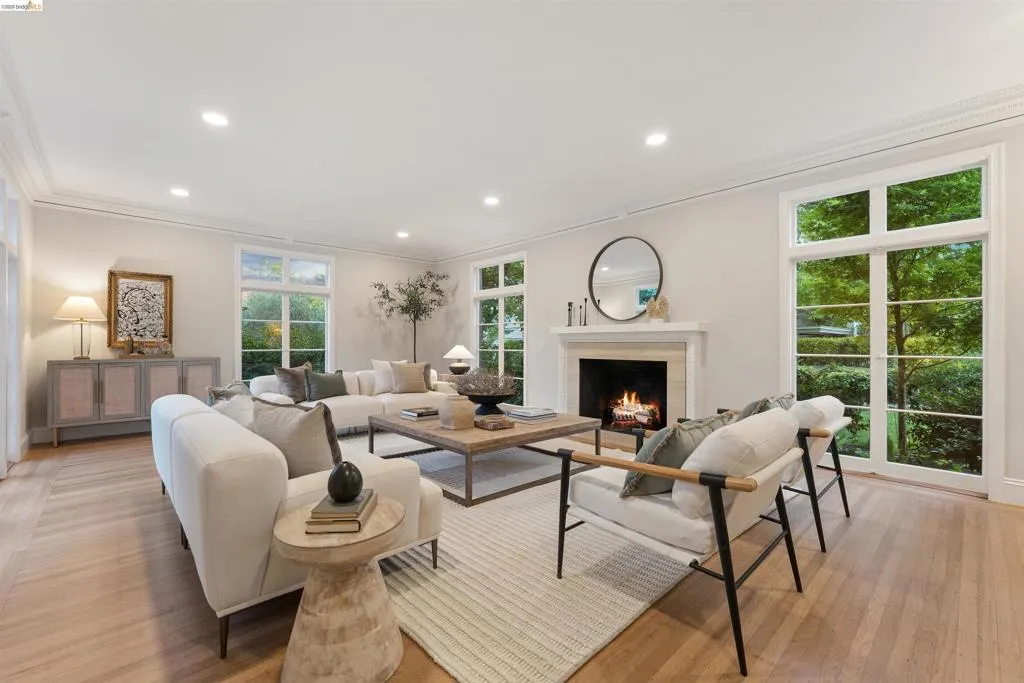 Spacious living area featuring light wood-finish flooring, a white mantel fireplace, abundant natural light from multiple paneled windows, recessed ceiling lighting, and crown molding. Fire has been photoshopped by Open Homes Photography.