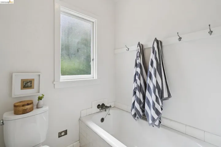 Bathroom featuring a white bathtub with tile surround, a privacy window, wall-mounted towel hooks, and a white toilet