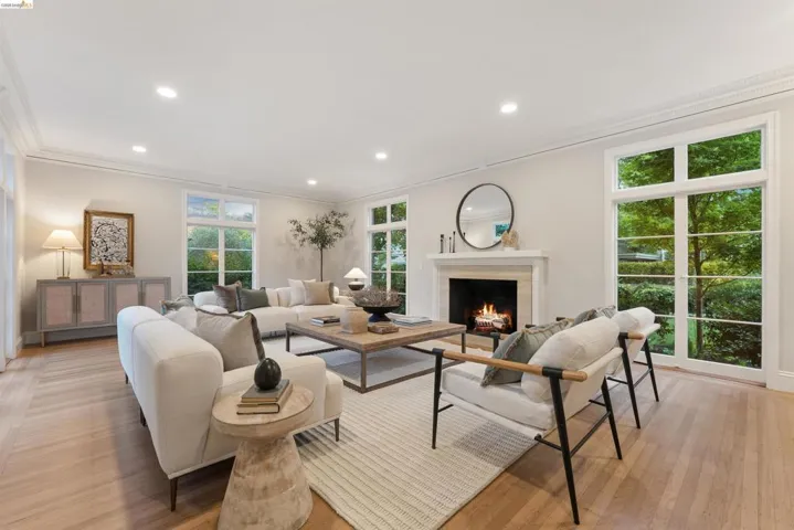 Spacious living area featuring light wood-finish flooring, a white mantel fireplace, abundant natural light from multiple paneled windows, recessed ceiling lighting, and crown molding. Fire has been photoshopped by Open Homes Photography.