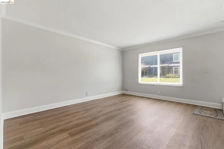 Spacious room featuring wood-finish flooring, white baseboards, crown molding, and a large window