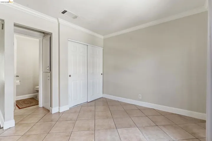 Neutral-toned tile flooring throughout a room featuring crown molding, white baseboards, and a built-in closet with white bifold doors