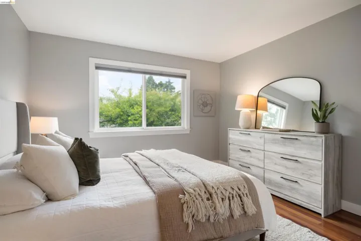 Hardwood-style flooring in a light-filled bedroom featuring a large window with white trim, a light grey upholstered headboard, and light grey wall paint